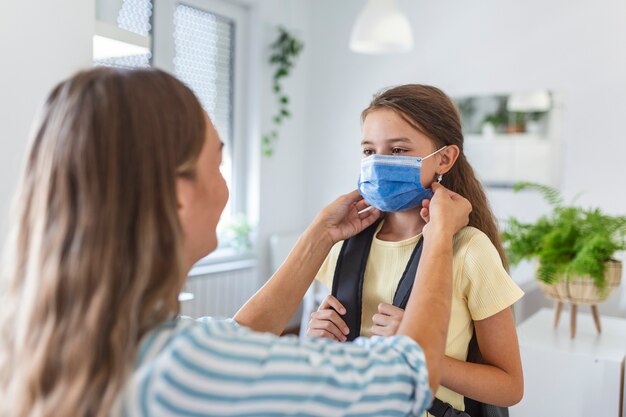 La giovane madre aiuta sua figlia a indossare una maschera medica per prepararsi ad andare a scuola. Evitare l'epidemia di Covid-19 o coronavirus. Di nuovo a scuola