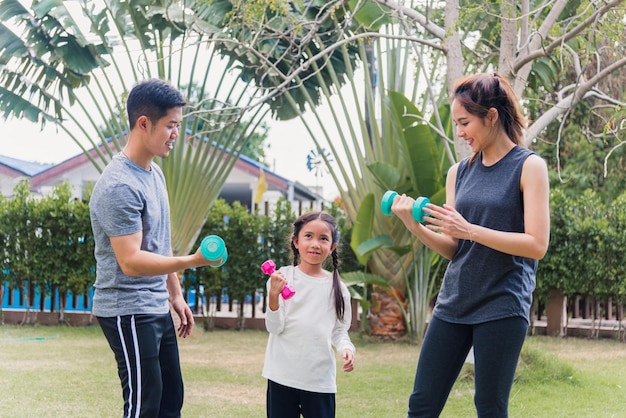 La giovane madre asiatica, il padre e la figlia del bambino che fanno esercizio insieme ai manubri è divertente all'aperto nella natura di un parco giardino di campo. Happy family kid sport ed esercizi per uno stile di vita sano