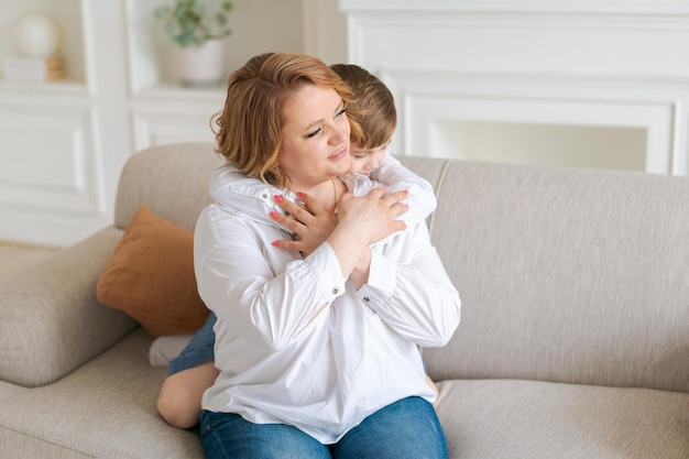 La giovane madre caucasica felice abbraccia il piccolo figlio in età prescolare insieme a casa seduto sul divano in brig...