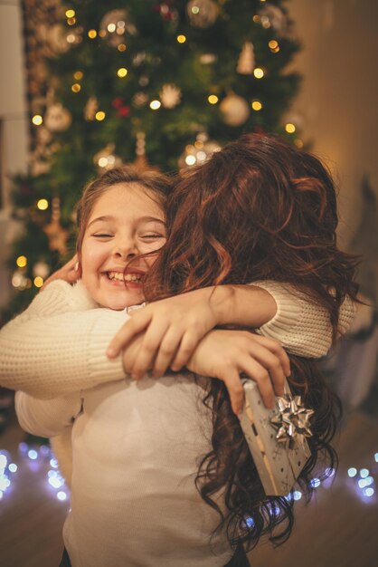 La giovane madre e la sua piccola figlia carina si abbracciano e si divertono accanto all'albero di Natale a casa.