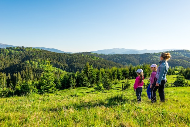 La giovane madre e le due figlie piccole viaggiatrici stanno su un pendio con una splendida vista sulle colline ricoperte da una fitta foresta di abeti contro il cielo blu in una calda giornata estiva. Concetto di turismo familiare