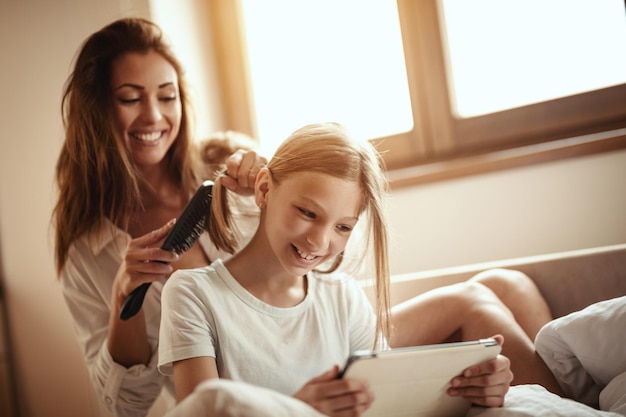 La giovane madre felice sorridente sta pettinando i capelli della sua piccola figlia sul letto mentre la ragazza sta guardando la tavoletta digitale.