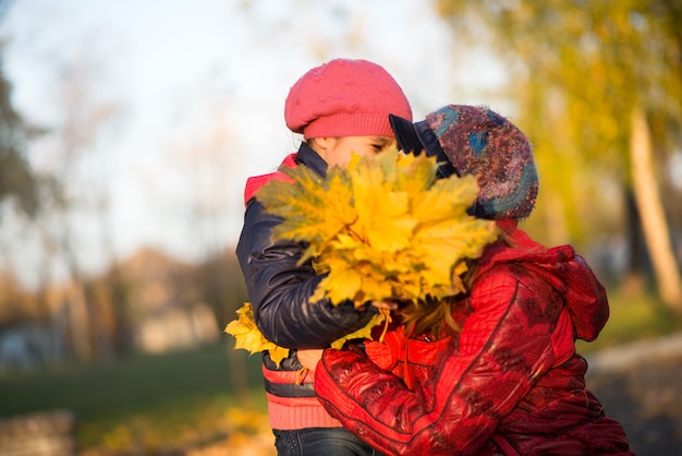 La giovane madre premurosa bacia e abbraccia la sua piccola figlia mentre cammina nel parco d'autunno con un bouquet di foglie d'acero gialle in una giornata calda e soleggiata. Concetto di amore e cura della famiglia