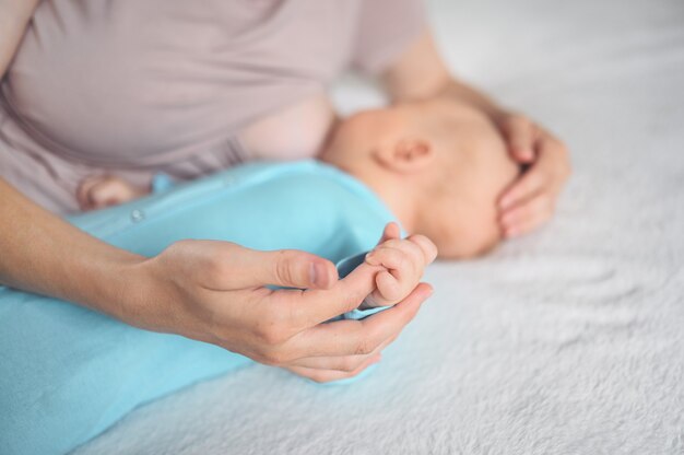 La giovane madre si trova con un neonato neonato carino in tuta blu sul letto, abbraccialo e lo allatta con il latte materno.