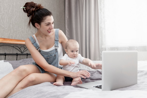 La giovane madre sorridente felice con un bambino piccolo lavora da casa, sedendosi sul letto.