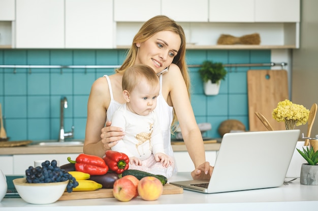 La giovane madre sta cucinando e giocando con sua figlia in una cucina moderna. Guardando il portatile, sorridendo.