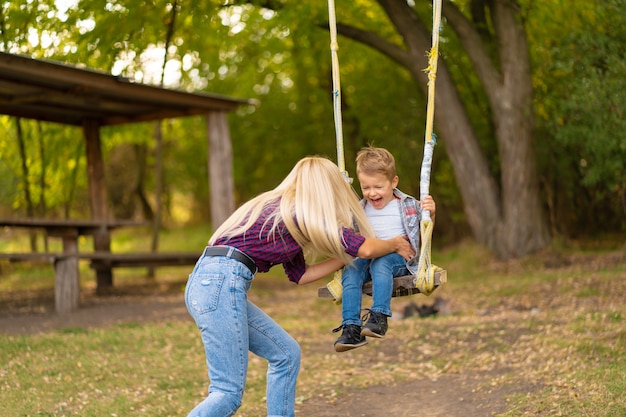 La giovane mamma bionda scuote il suo piccolo figlio su un'altalena in un parco verde. Infanzia felice.