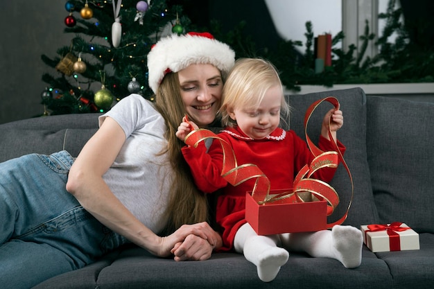 La giovane mamma con il cappello di Babbo Natale e il bambino disimballano il regalo di Natale. Capodanno.