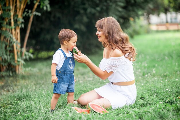 La giovane mamma e il figlio piccolo hanno giocato sull'erba. Mamma e figlio felici si siedono sull'erba e mangiano l'anguria. Estate.