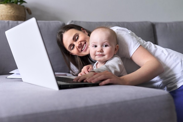 La giovane mamma sorridente con il bambino guarda insieme il monitor del laptop seduto a casa sul divano