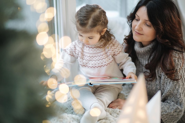 La giovane nonna gioca e legge un libro con il suo adorabile grandaugher nea abete natalizio