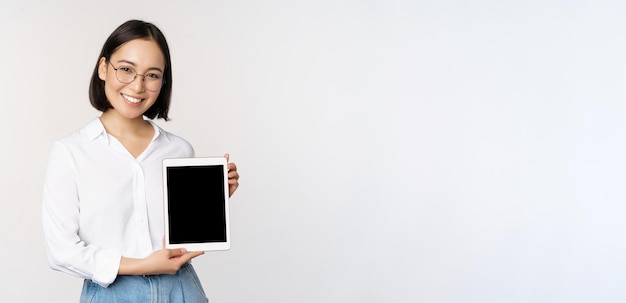La giovane professionista asiatica mostra le informazioni sullo schermo del tablet digitale sul suo gadget sorridendo e guardando la fotocamera in piedi su sfondo bianco