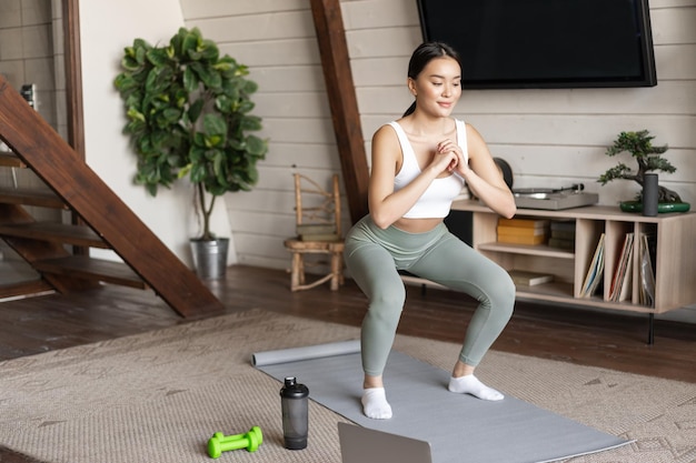 La giovane ragazza asiatica di forma fisica che fa l'allenamento di squat a casa segue l'istruttore di palestra di sport online in piedi su...