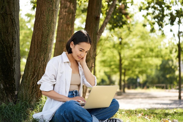 La giovane ragazza asiatica felice si siede nel parco vicino all'albero guardando il computer portatile che lavora in remoto da talki all'aperto