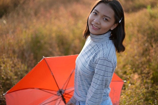 La giovane ragazza asiatica tiene l'ombrello arancione e sorride all'erba archiviata marrone.
