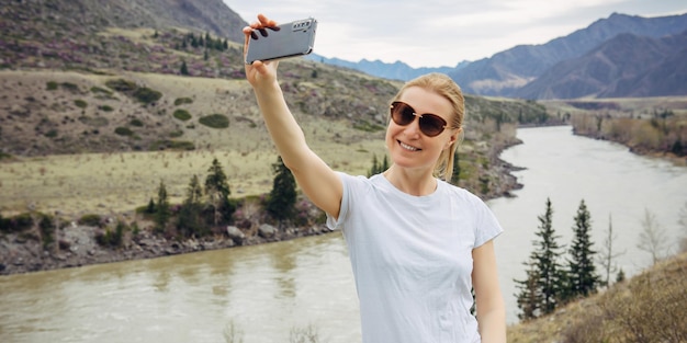 La giovane ragazza bionda in occhiali da sole e maglietta bianca prende un selfie sul suo smartphone sullo sfondo del fiume e delle montagne rocciose. Turista felice sorride alla fotocamera del telefono cellulare.