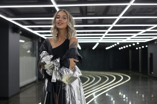 La giovane ragazza bionda in un mantello d'argento brillante e con un trucco interessante sta al chiuso con una foto di alta qualità del soffitto leggero ceiling