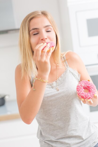 La giovane ragazza bionda mangia le ciambelle rosa nella cucina di casa con emozioni di gusto.