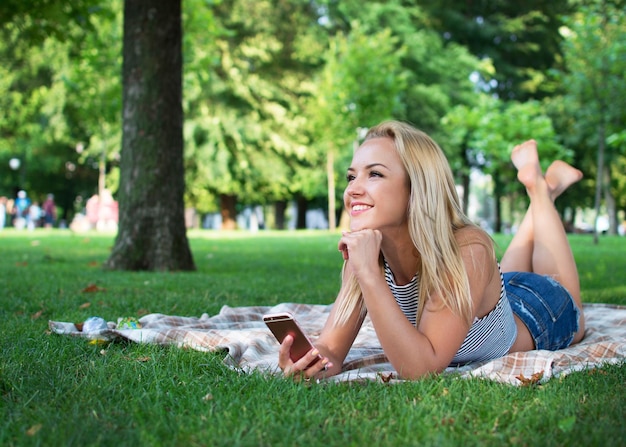 La giovane ragazza bionda molto bella è sdraiata sull'erba in una soleggiata giornata estiva facendo un selfie con il telefono