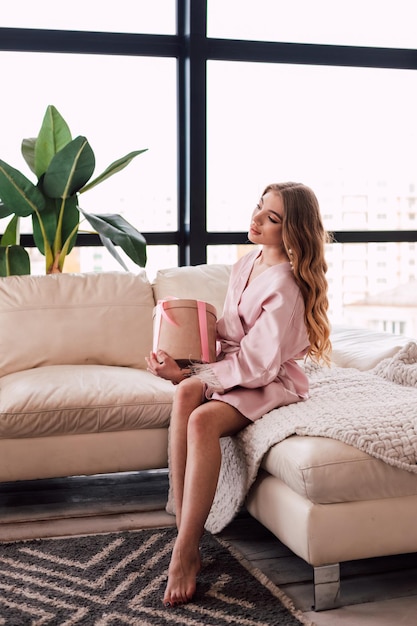 La giovane ragazza bionda sexy felice con le gambe lunghe di mattina apre un regalo che guarda fuori dalla finestra