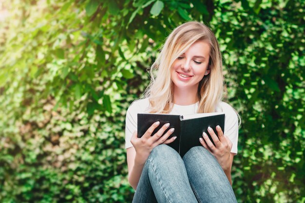 La giovane ragazza bionda si siede su una panchina, legge un libro e sorride in un parco su uno sfondo di alberi e cespugli