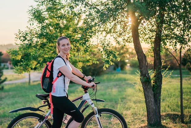 La giovane ragazza bionda sportiva in vestiti di sport con uno zaino guida una bici e sorride alla macchina fotografica