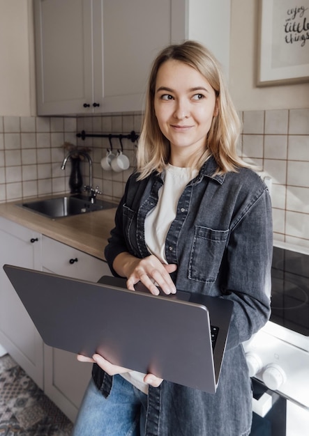 La giovane ragazza bionda sta con un computer portatile in cucina e lavora da casa