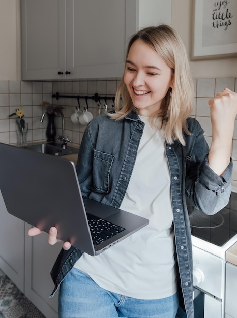 La giovane ragazza bionda sta con un computer portatile in cucina e lavora da casa