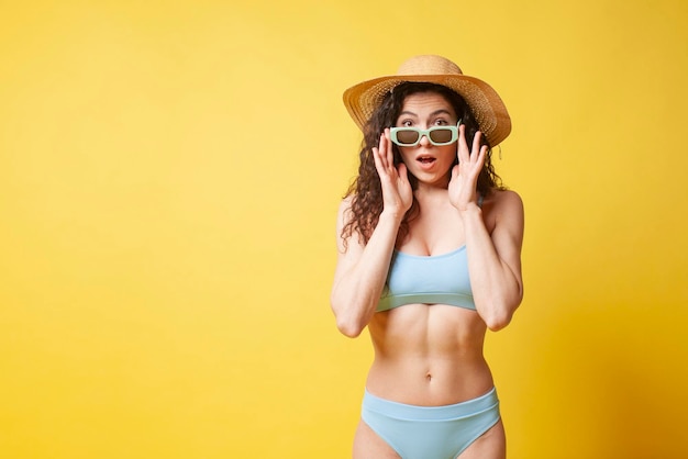 La giovane ragazza castana scioccata in costume da bagno blu e con i capelli ricci tiene gli occhiali da sole