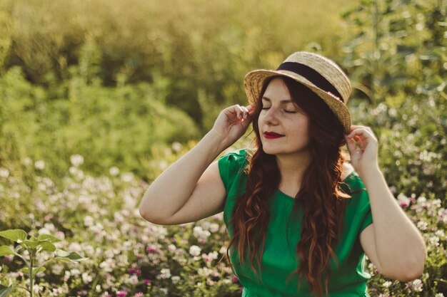 La giovane ragazza caucasica in un vestito verde e un cappello di paglia sorride in un giacimento di fiore un giorno soleggiato.