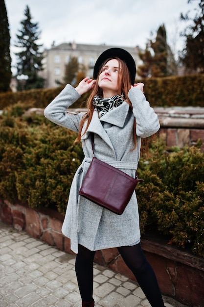 La giovane ragazza di modello in un cappotto grigio e un cappello nero con la borsa di cuoio sulle spalle ha posato vicino ai cespugli alla via della città.
