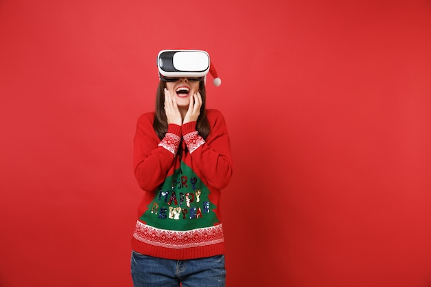 La giovane ragazza di Santa stupita in maglione lavorato a maglia, cappello di Natale che guarda in cuffia, mette le mani sul viso isolato su sfondo rosso. Felice Anno Nuovo 2019 celebrazione festa concetto. Mock up copia spazio.