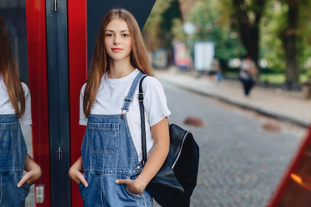 La giovane ragazza graziosa con la cartella cammina sulle riflessioni urbane della via