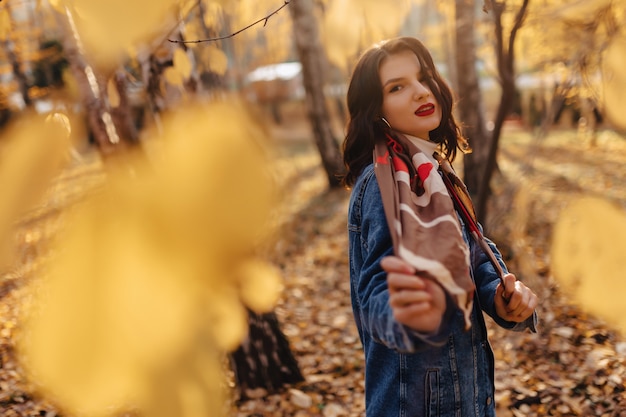 La giovane ragazza graziosa in un cappotto del denim cammina nel parco di autunno