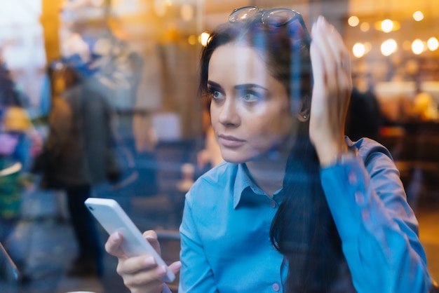 La giovane ragazza impegnata in una camicia blu è seduta in un caffè, tiene in mano uno smartphone