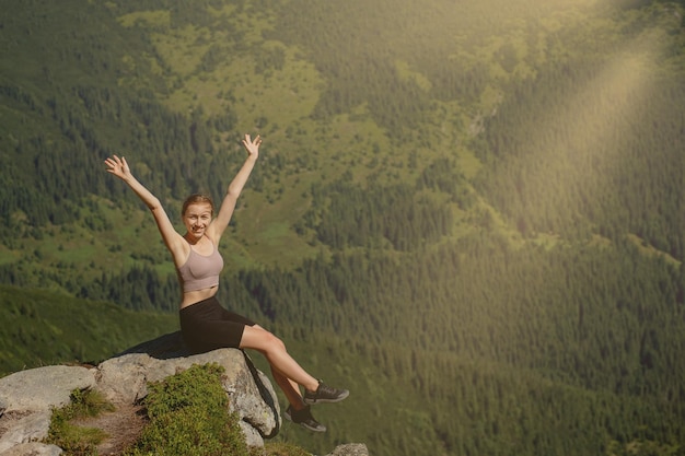 La giovane ragazza seduta in cima alla montagna ha alzato le mani sullo sfondo della foresta La donna è salita in cima e si è goduta il successo