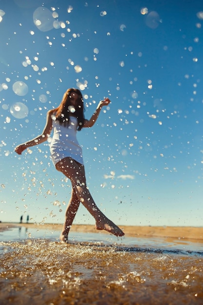 La giovane ragazza snella sfocata in un vestito bianco corre sull'acqua con schizzi nel concetto di luce solare