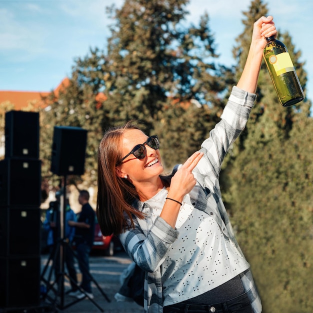 La giovane ragazza sorridente che indossa occhiali da sole tiene una bottiglia di vino. Stile casual da donna. Persona di sesso femminile che si diverte. Buone feste di festa. Ritratto di stile di vita all'aperto. Bella faccia da adulto. Evento di strada