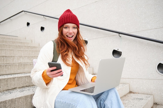 La giovane ragazza turistica che prenota una camera d'albergo sul laptop inserisce il codice di conferma sullo smartphone seduto con