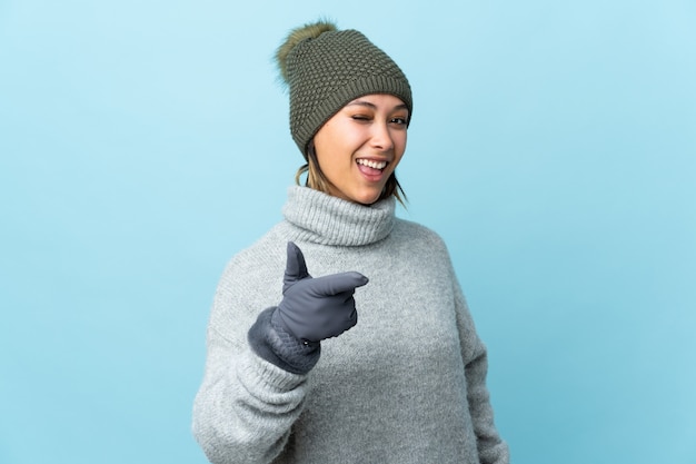 La giovane ragazza uruguaiana con il cappello dell'inverno isolato sulla parete blu indica il dito voi