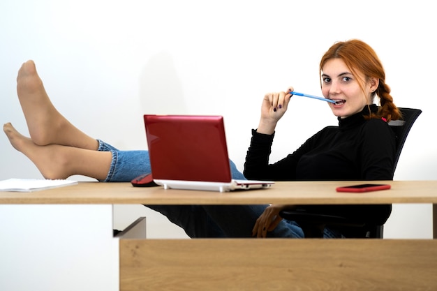 La giovane seduta felice della donna di impiegato si è distesa con i piedi sulla tabella dietro lo scrittorio funzionante con il computer portatile, il telefono cellulare ed il taccuino.