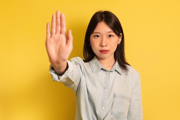 La giovane signora asiatica che gesturing smette di mostrare il palmo alla macchina fotografica su sfondo giallo per studio