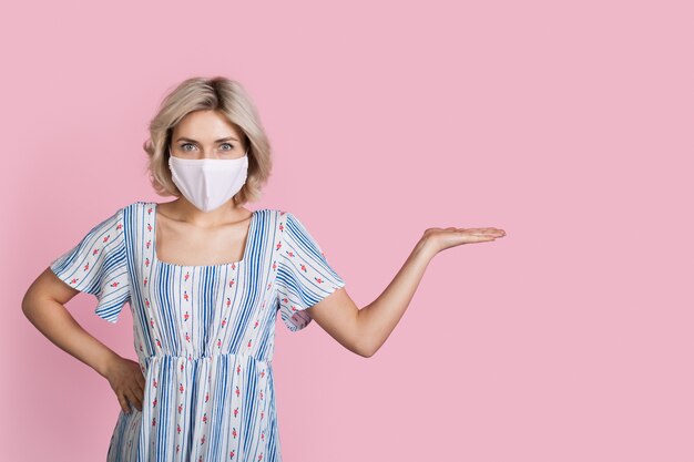 La giovane signora bionda in un vestito che porta una mascherina medica sta pubblicizzando qualcosa su una tenuta rosa della parete è con la palma