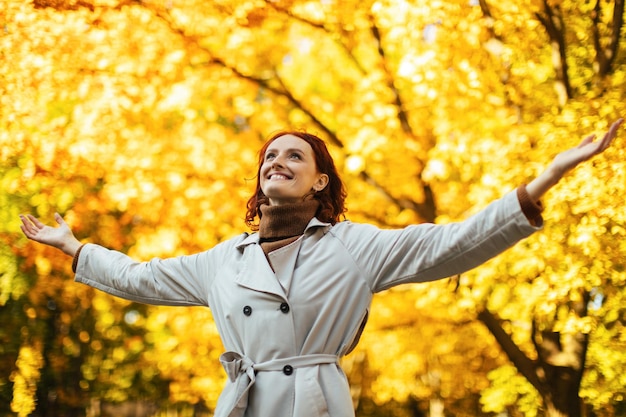 La giovane signora europea sorridente in impermeabile gode della libertà del bel tempo nel tempo libero nel parco con le foglie gialle