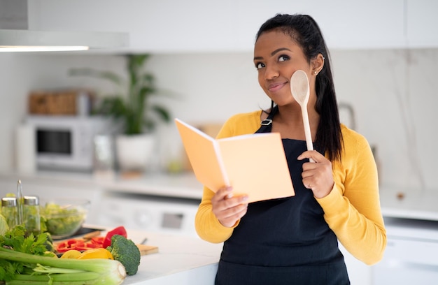 La giovane signora nera sorridente e pensierosa cuoca in grembiule con il cucchiaio di legno legge il libro di ricette pensa al nuovo piatto