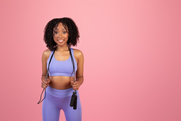 La giovane signora riccia afroamericana sorridente in abbigliamento sportivo tiene la corda per saltare si diverte a fare sport da sola