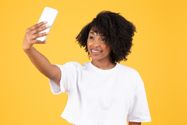 La giovane signora riccia nera allegra in maglietta bianca fa il selfie sullo smartphone per il blog di social media