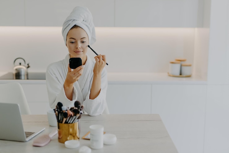 Foto la giovane signora rinfrescata applica la cipria con il pennello cosmetico si guarda in pose speculari in cucina a casa si siede alla scrivania con prodotti di bellezza indossa un asciugamano avvolto e una vestaglia bianca