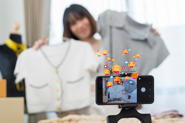 La giovane stilista asiatica che usa il telefono cellulare riceve l'ordine di acquisto e mostra i vestiti che registrano il video in diretta streaming online nel negozio