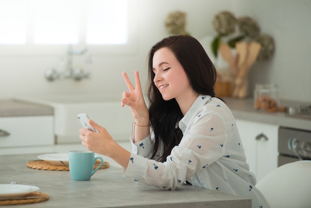 La giovane studentessa bruna è seduta in cucina con lo smartphone mobile